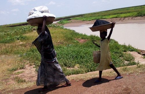 UNa mujer y una niña, con grandes fardos en la cabeza caminan por una carretera de Sudán